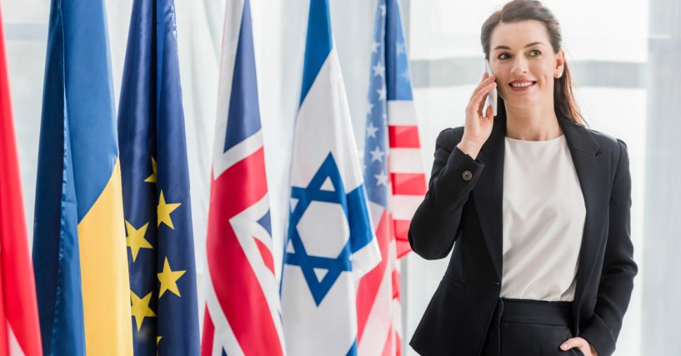 Executiva sorridente ao telefone em frente a bandeiras de diversos países, ilustrando a comunicação internacional corporativa.