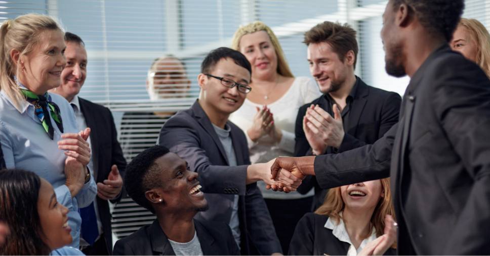 Grupo multiétnico aplaude cumprimento entre colaboradores em ambiente de comunicação intercultural.