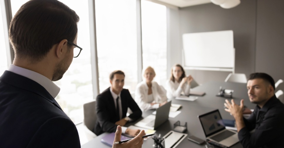 Colaboradores sentados ao redor da mesa escutam o palestrante durante a reunião corporativa em inglês.