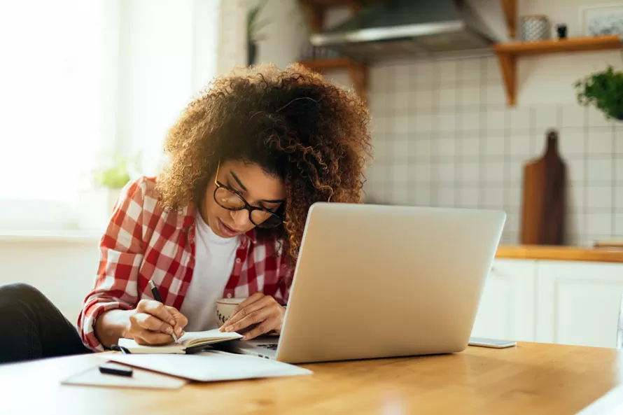 mulher anotando estratégias de estudo no papel após pesquisar no notebook